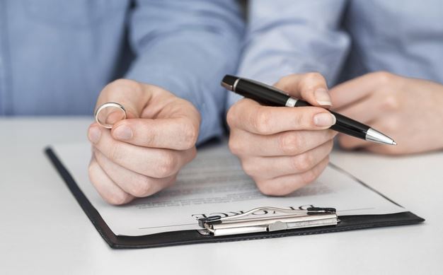 One hand holding a ring and another hand holding a pen over a divorce document.