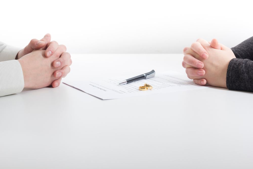 Two hands folded across from each other with a paper, pen, and two wedding bands between them